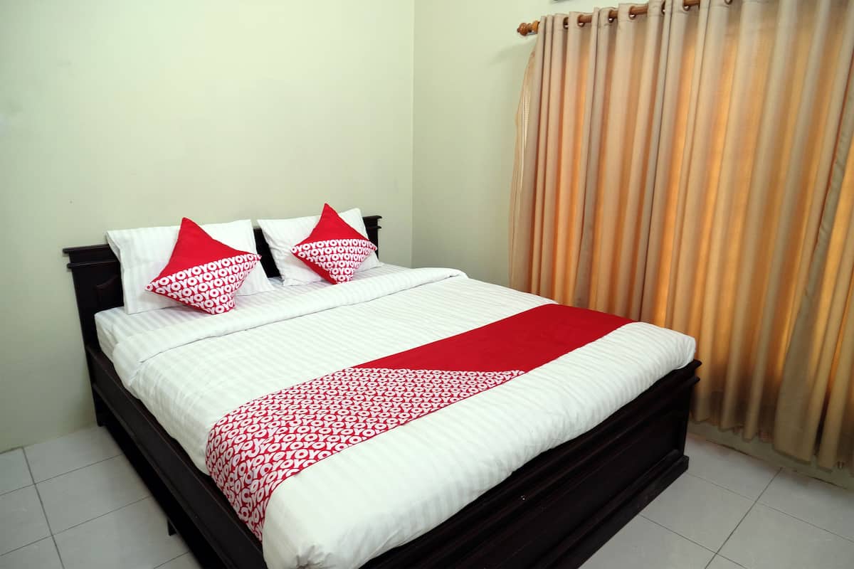 A neatly made bed with white bedding and red accents, a dark wooden headboard, and a beige curtain.