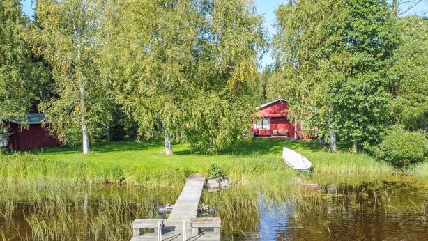 Une jetée en bois s’étendant sur une étendue d’eau calme, entourée d’une végétation luxuriante et avec un bâtiment rouge en arrière-plan.