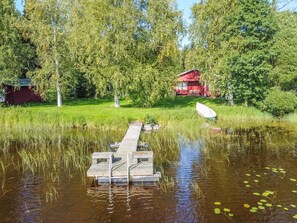 Une jetée en bois s’étendant sur une étendue d’eau calme, entourée d’une végétation luxuriante et avec un bâtiment rouge en arrière-plan.