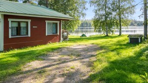 Une cabane rouge avec un toit en métal, un chemin de gravier menant à celle-ci, et un lac en arrière-plan.