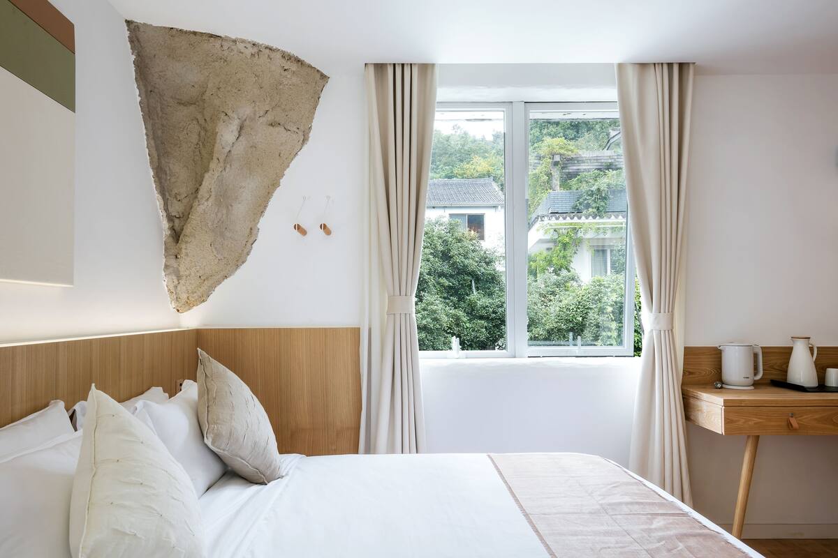 Un dormitorio con una cama grande, un cabecero de madera, una ventana con cortinas y un escritorio de madera con una tetera y tazas.