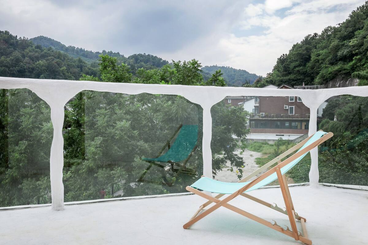 Un balcón con barandal blanco, una silla azul y vistas a un tobogán verde y edificios al fondo.