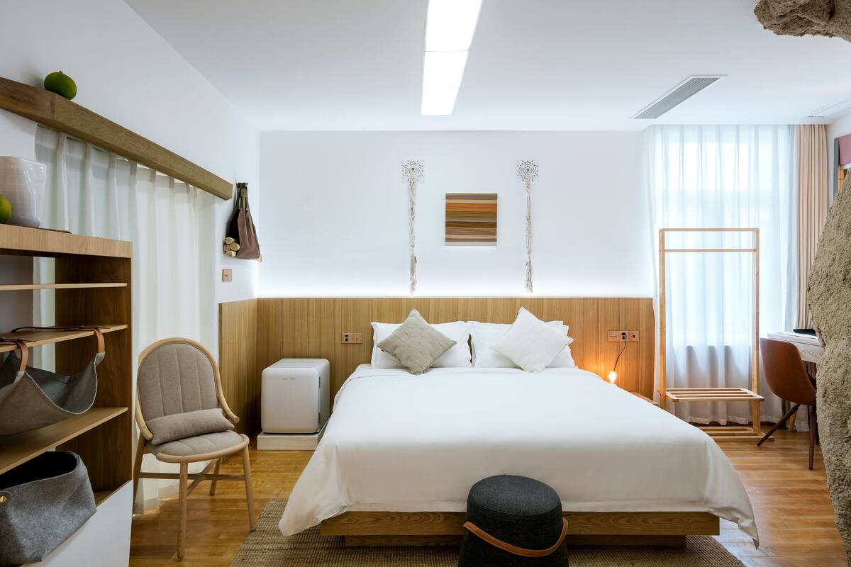 Una habitación de hotel moderna con cabecera de madera, cama con ropa blanca, una silla, una mesita y un escritorio.
