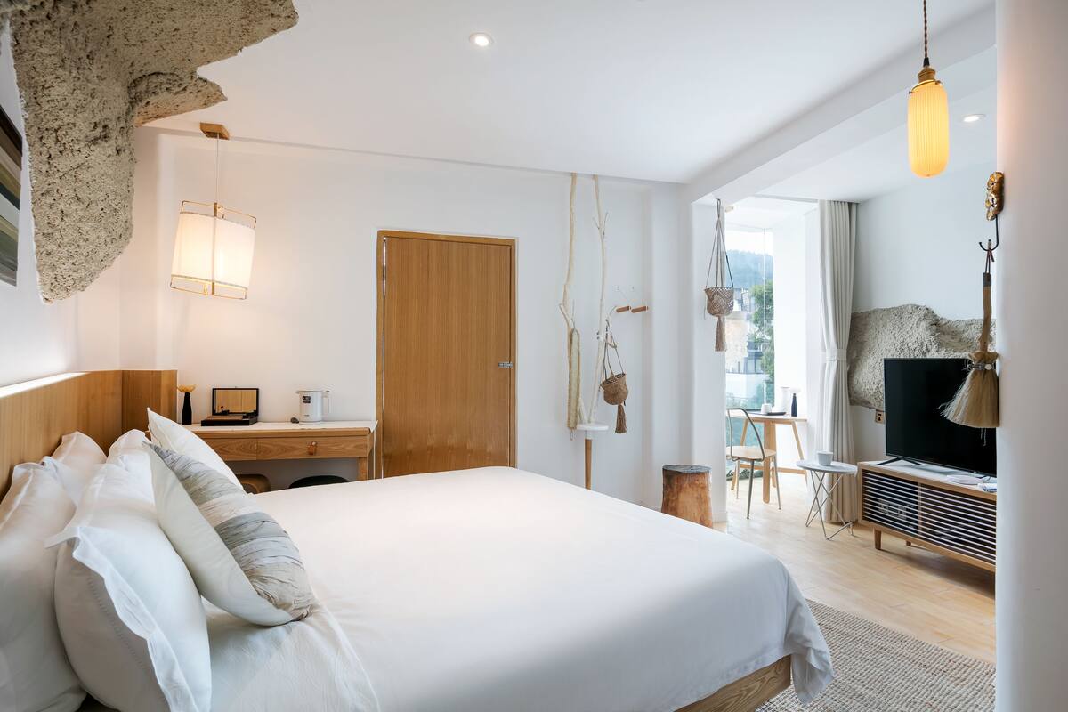 Una habitación de hotel moderna con una cama grande, cabecera de madera, un escritorio con computadora, televisor y vista al exterior.