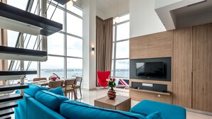 Ein modernes Wohnzimmer mit großem Fernseher, blauem Sofa und Blick auf die Stadt durch bodentiefe Fenster.