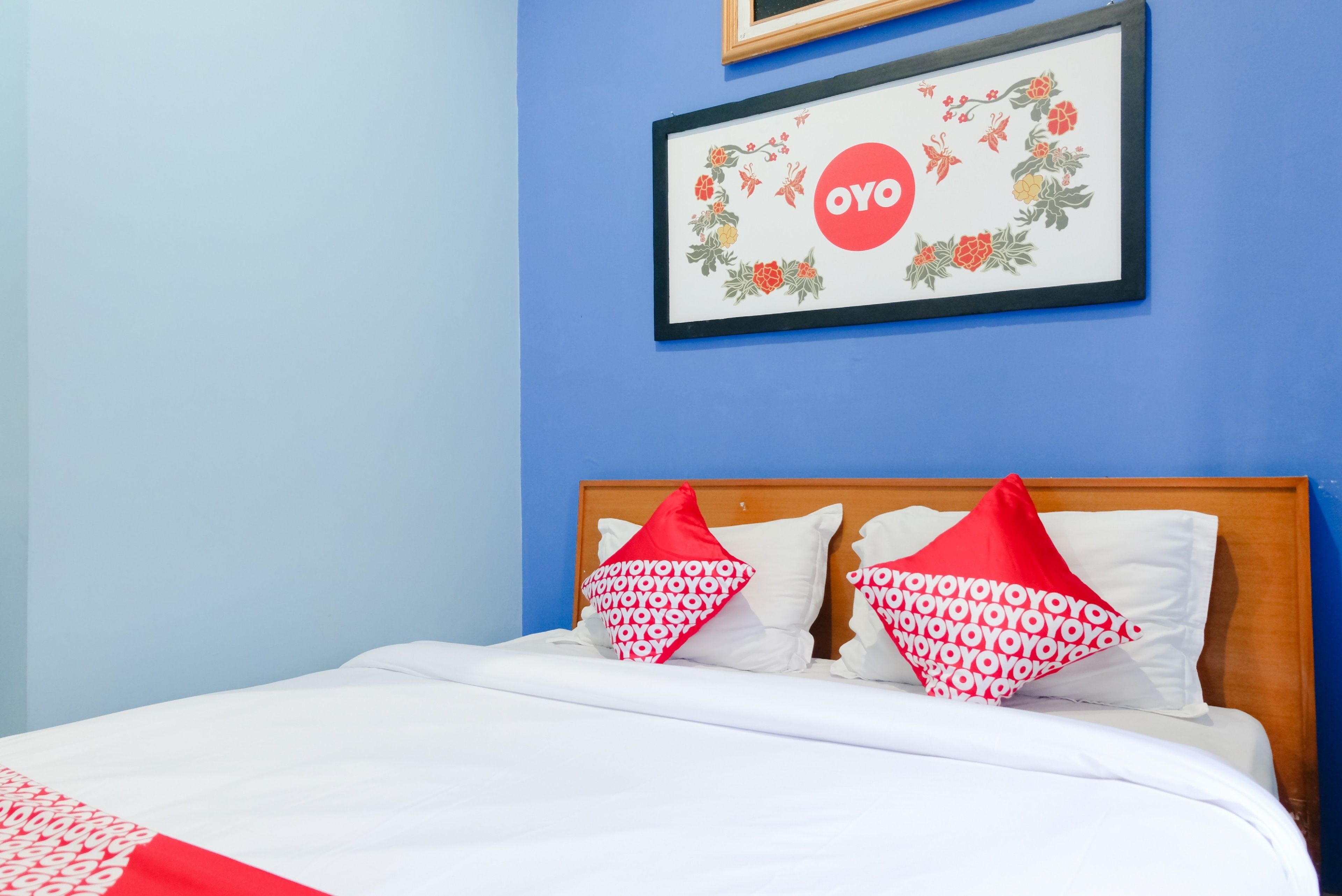 A neatly made bed with white bedding and red pillows, a framed artwork with floral designs and the word "OYO" on the wall, and a blue wall in the background.