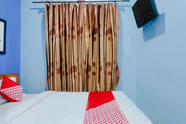 A hotel room with a bed, a red pillow, a curtain with a floral pattern, and a wall-mounted TV.