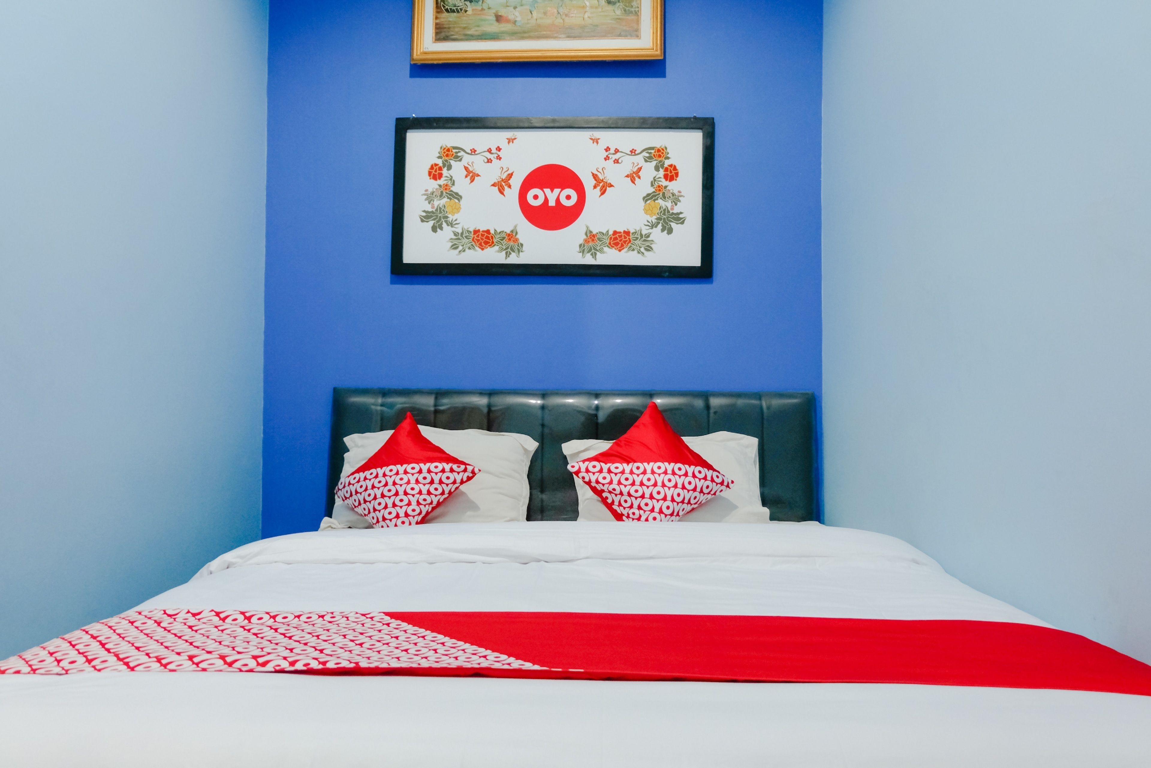 A bedroom with a blue wall, a framed artwork with 'OYO' logo, a bed with white and red bedding, and a red throw blanket.