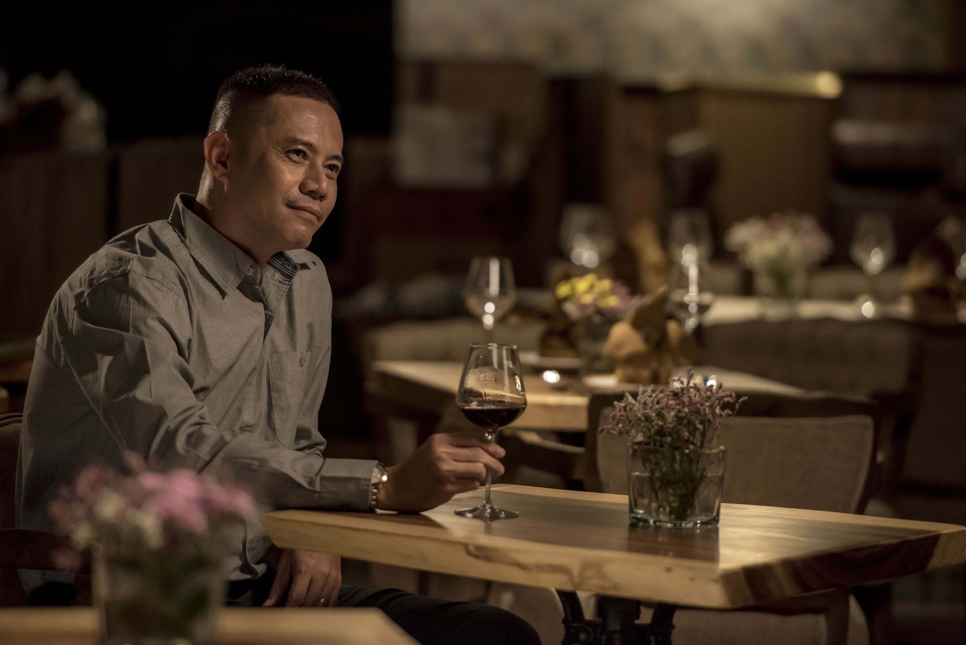 Un hombre sentado a una mesa de madera en un restaurante con poca luz, copas de vino y un jarrón de flores.