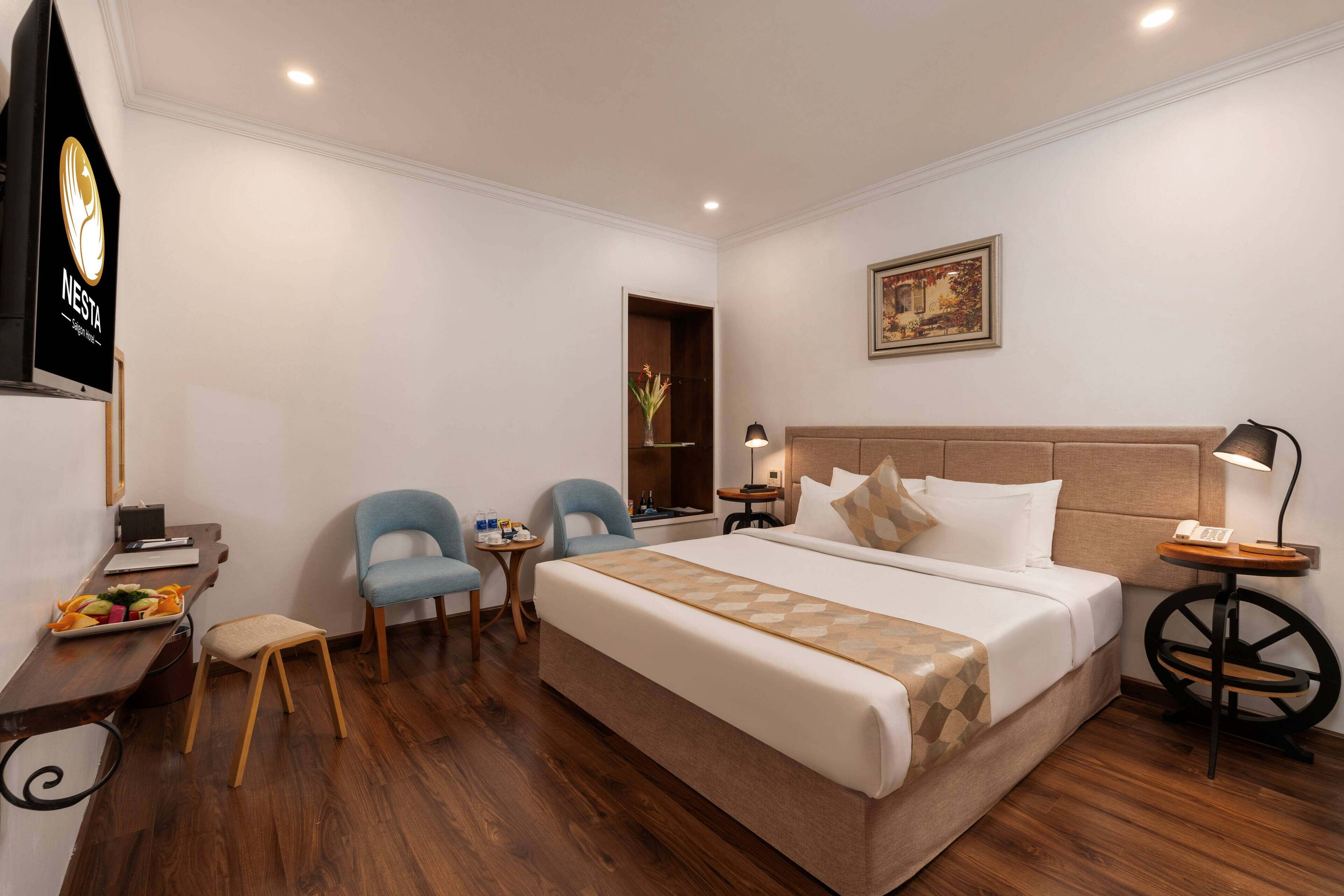 A hotel room with a large bed, two armchairs, a small table, a desk with a computer, a nightstand with a lamp, and a painting on the wall.