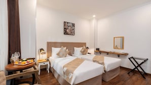 Quarto de hotel com duas camas, uma mesinha com uma bandeja de frutas e uma taça de vinho, mesa de cabeceira com abajur e um espelho na parede.