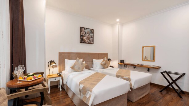 A hotel room with two beds, a small table with a tray of fruit and a glass of wine, a bedside table with a lamp, and a mirror on the wall. -  Nesta Hotel Saigon (Ho Chi Minh City)