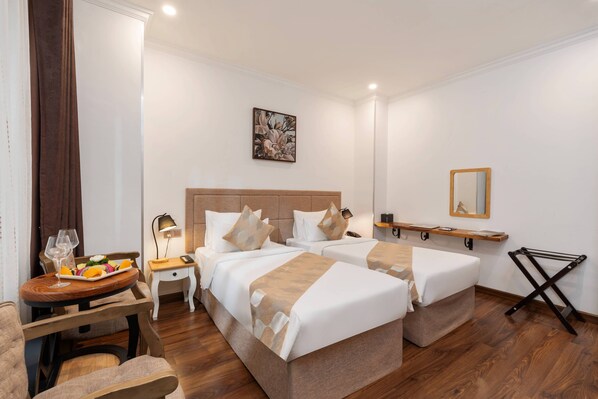 Een hotelkamer met twee bedden, een kleine tafel met een schaal fruit en een glas wijn, een nachtkastje met een lamp, en een spiegel aan de muur.