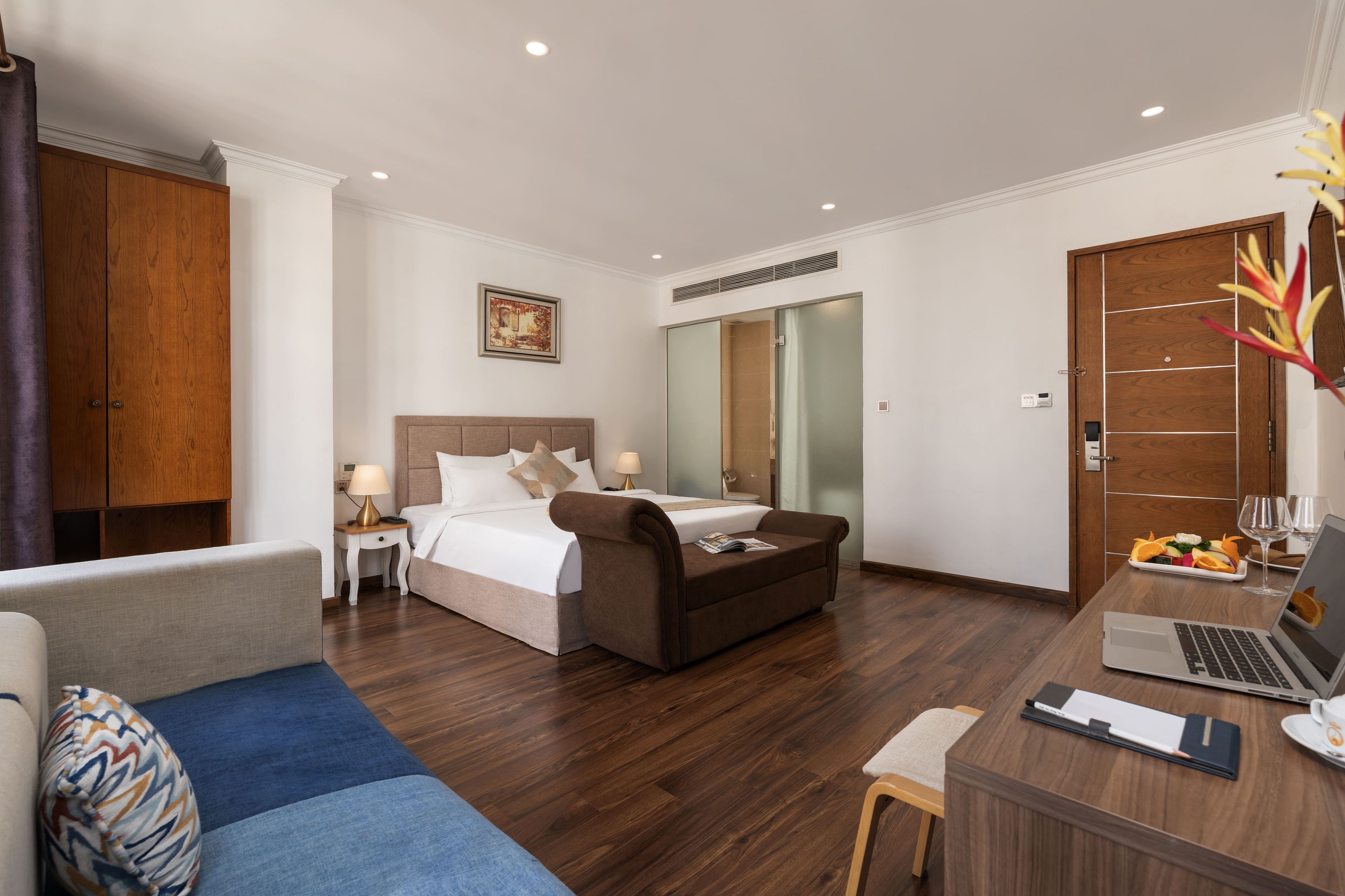 A hotel room with a bed, a desk with a laptop, a chair, a bedside table with a lamp, and a large mirror.