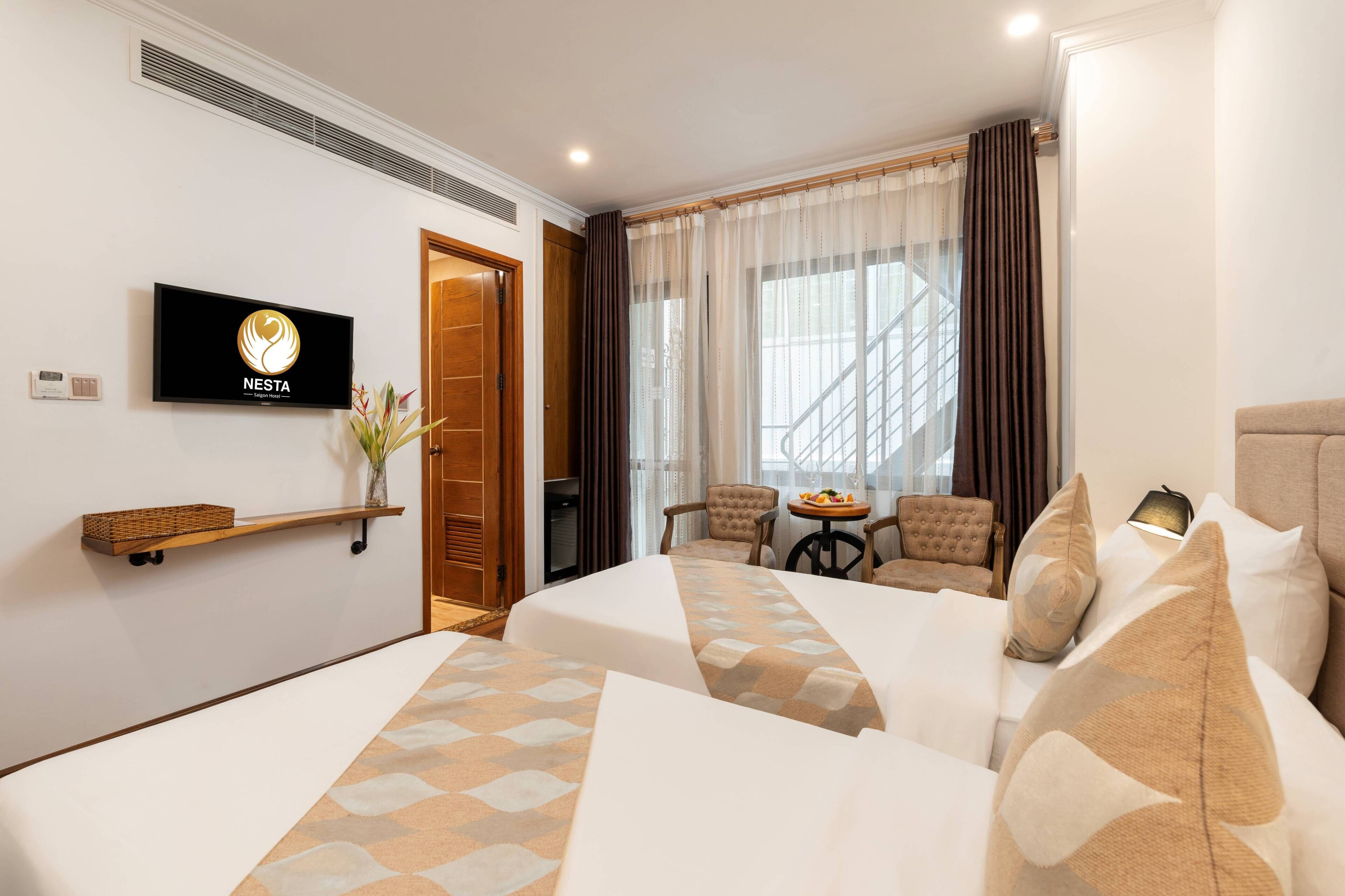 A hotel room with two beds, a TV, a small table with a vase, and a staircase visible through the window.