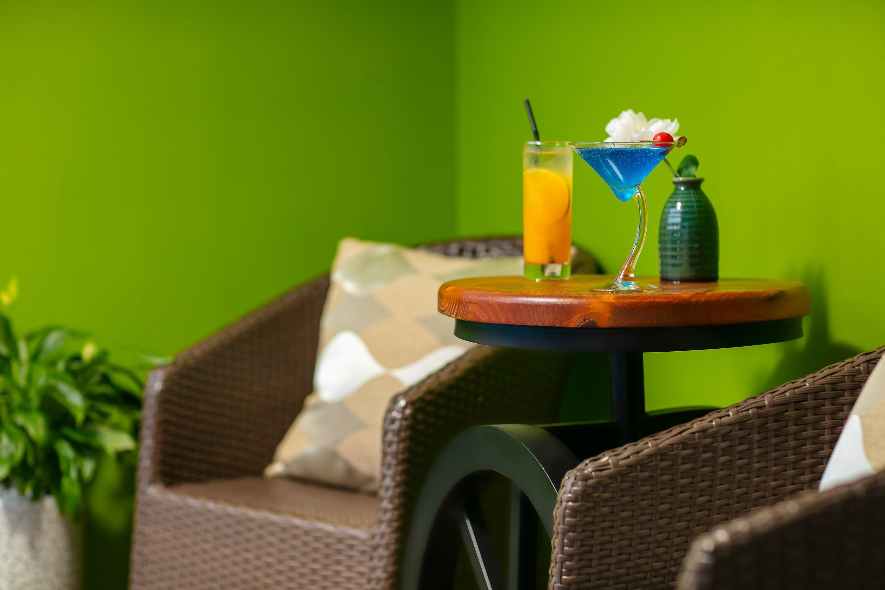 A wicker chair with a cushion, a small round table with a drink and a bottle, and a green wall in the background.