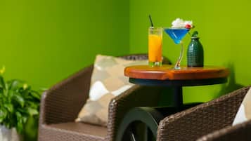 A wicker chair with a cushion, a small round table with a drink and a bottle, and a green wall in the background.