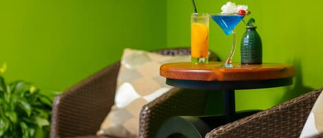 A wicker chair with a cushion, a small round table with a drink and a bottle, and a green wall in the background.