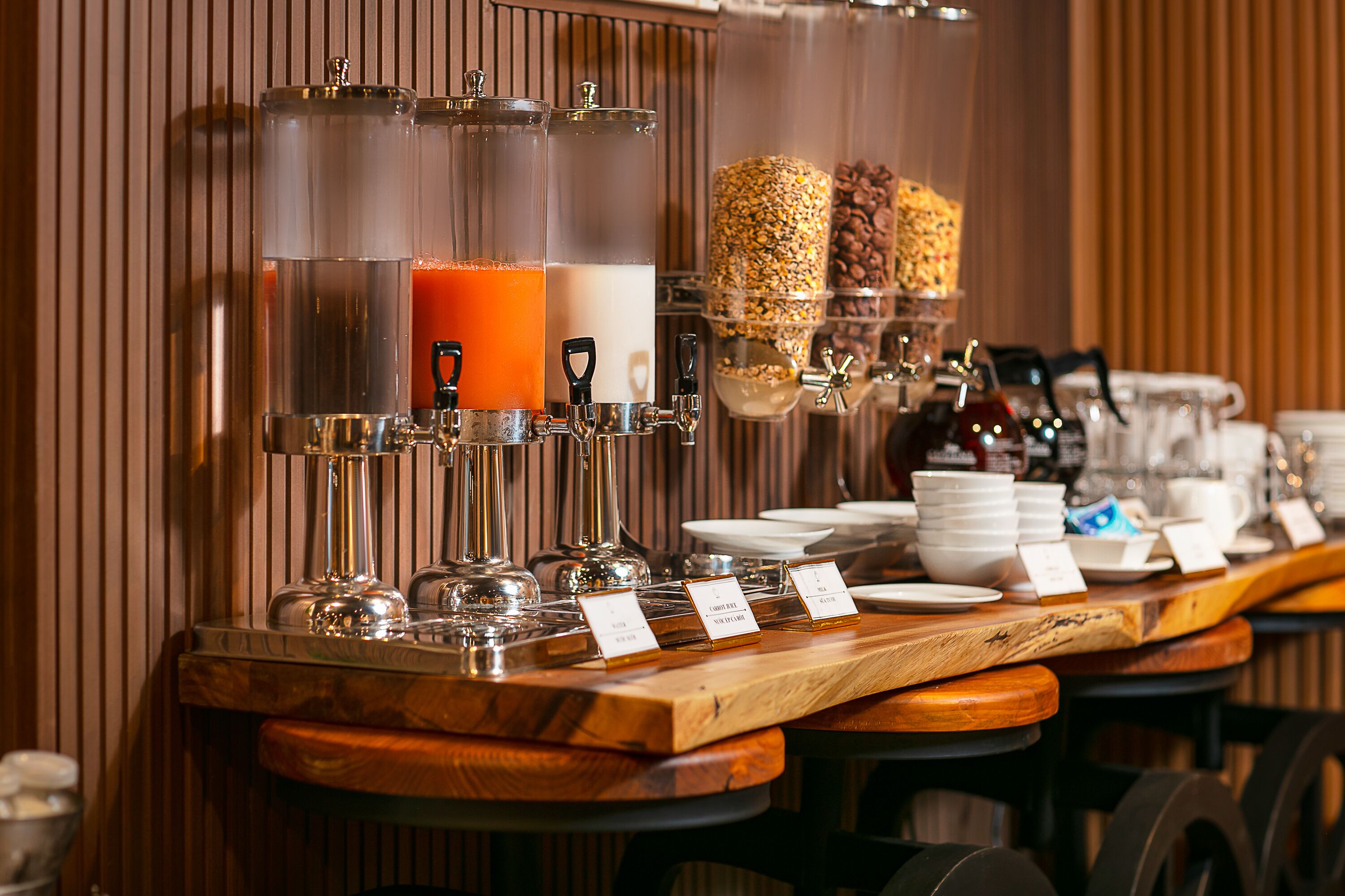 Ein Frühstücksbuffet mit verschiedenen Speisenspendern, darunter ein Saftspender und ein Getreidespender, mit einer Holztresen und Hockern.