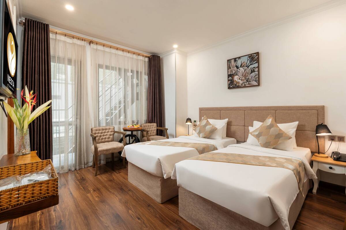 Een hotelkamer met twee bedden, een zithoek met een stoel en een tafel, een schilderij aan de muur en een groot raam met gordijnen.