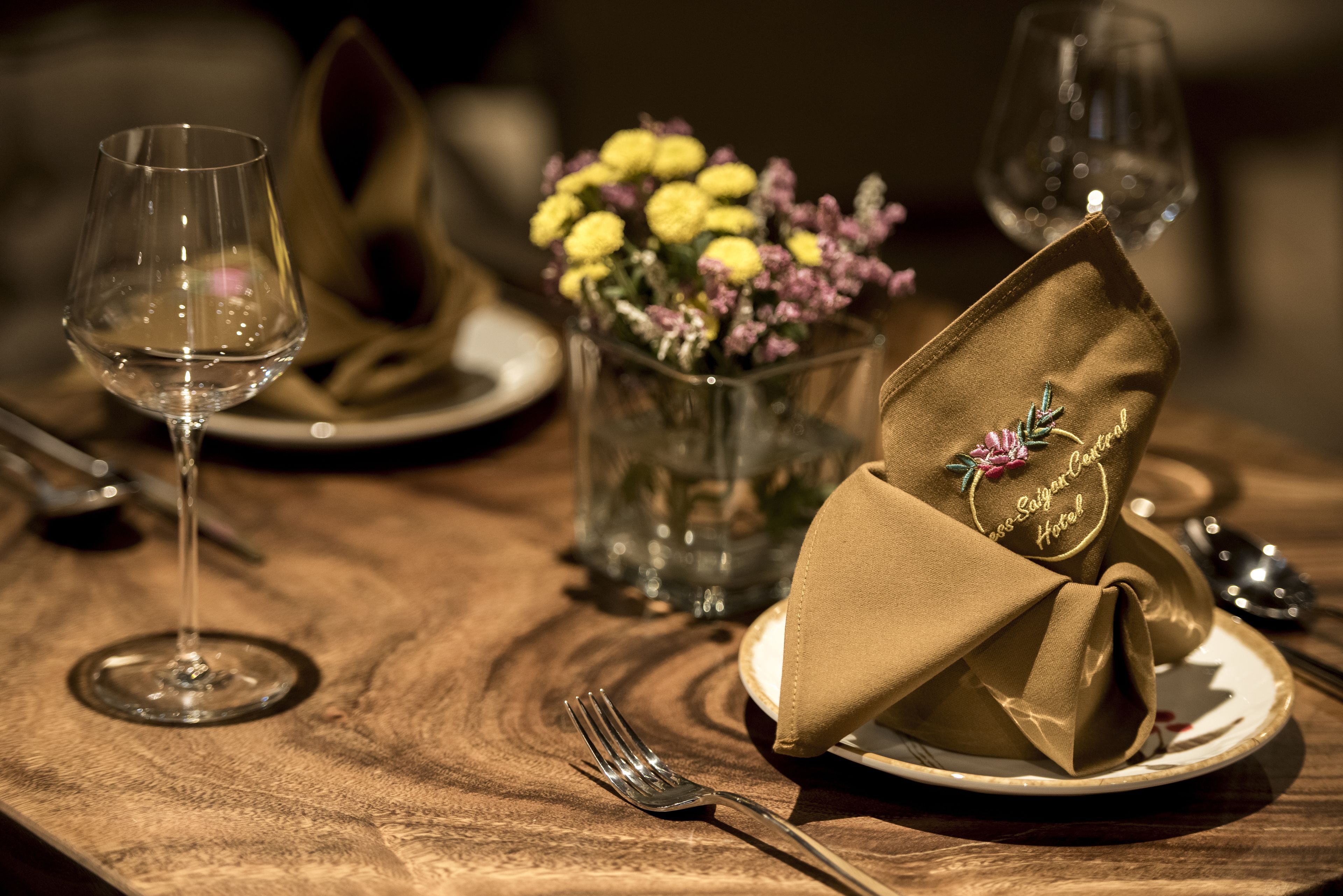 A dining table set with a wine glass, a folded napkin, a plate, and a vase with flowers.