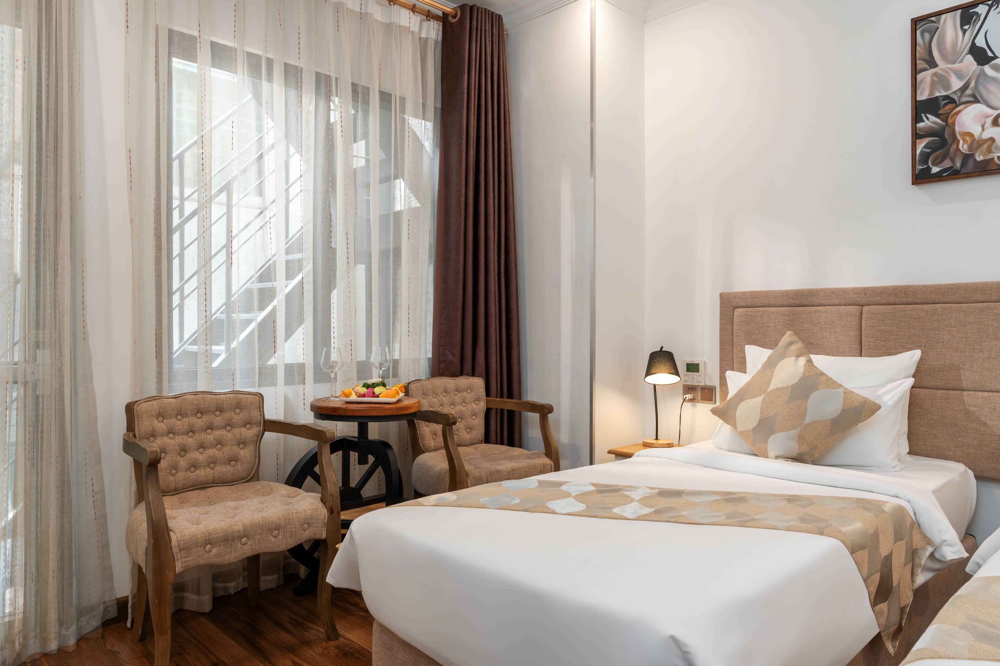 A hotel room with two beds, a small table with fruit, a chair, and a painting on the wall.