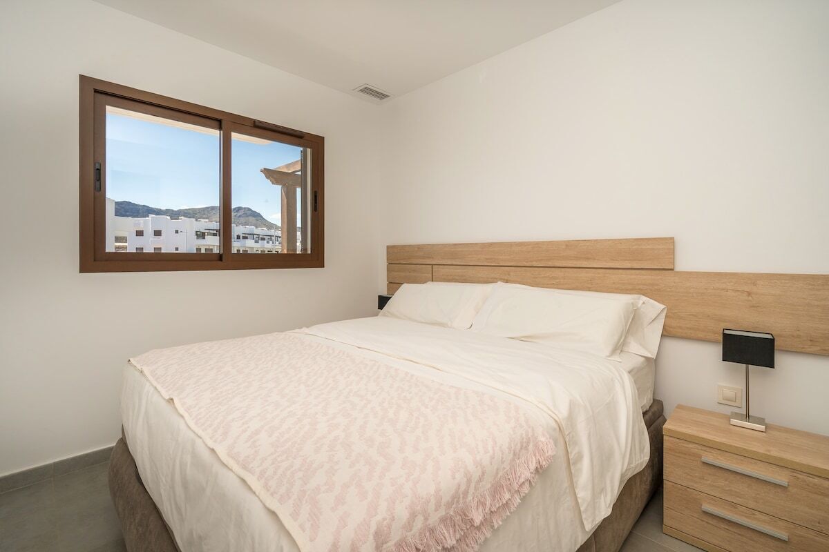 A bedroom with a bed, a nightstand, a window, and a view of buildings.