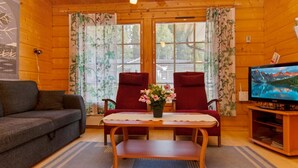 Een gezellig interieur van een chalet met een houten plafond, twee rode stoelen, een grijze bank, een houten salontafel met een bloevas, en een televisie op een houten dressoir.
