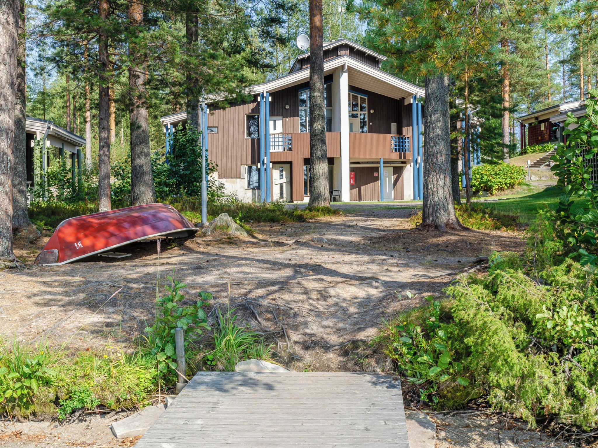 Un bâtiment à deux étages doté d’une terrasse en bois menant à l’entrée, entouré d’arbres et d’un bateau rouge.