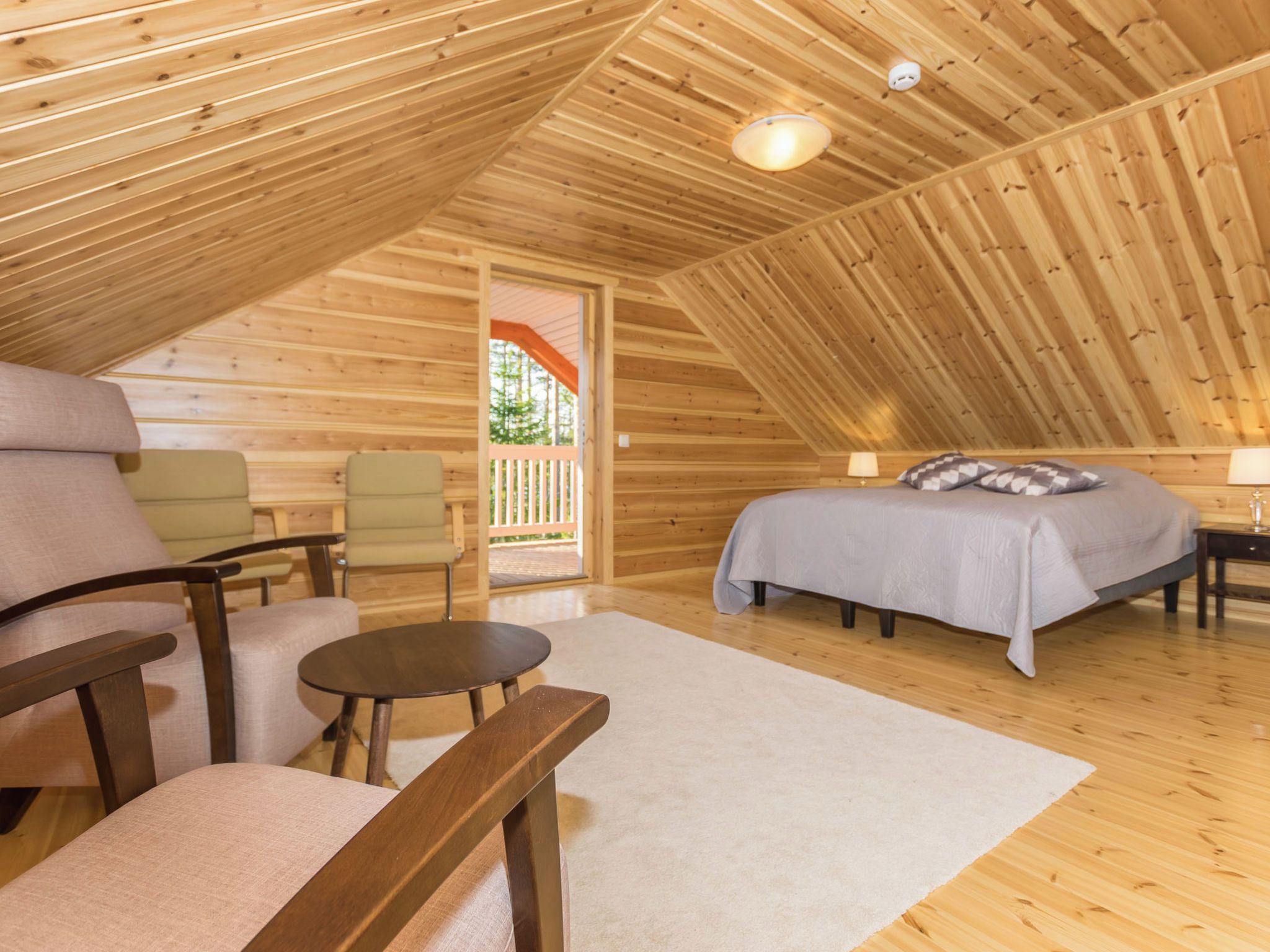 A cozy, wooden interior with a bed, two chairs, a small table, and a view of the outdoors.