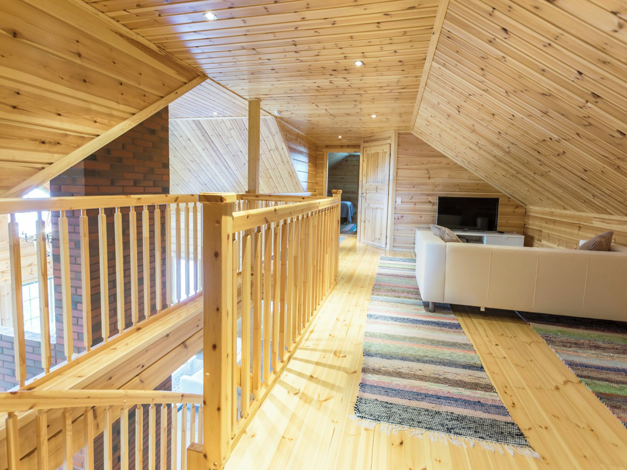 Ein geräumiges Dachzimmer mit Holzbalkendecke, einem weißen Sofa, einem Fernseher und einem farbenfrohen Teppich.