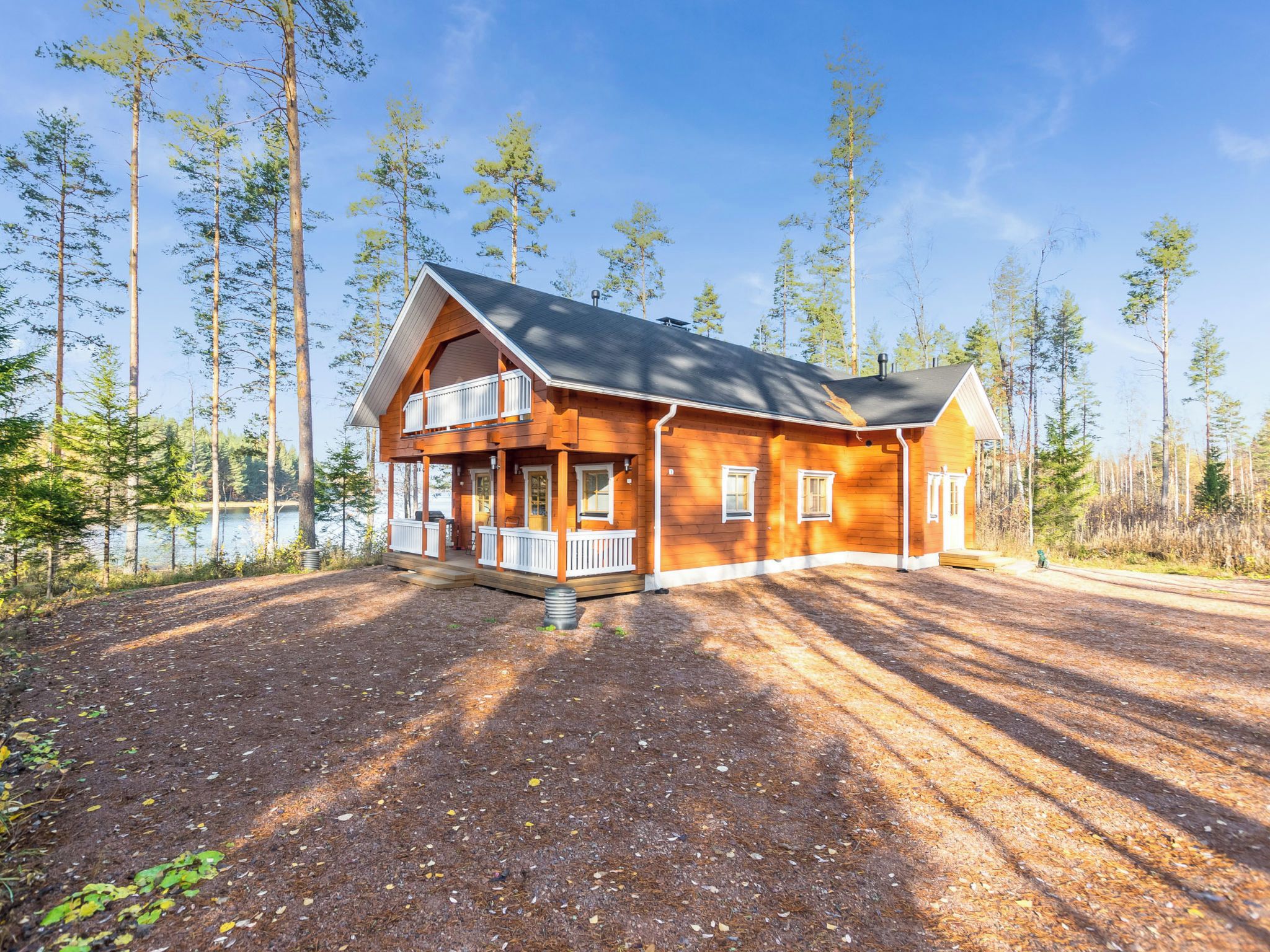 A two-story log cabin with a balcony, surrounded by a forest and a lake.