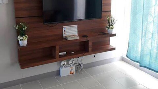 A modern living room with a large flat-screen TV mounted on a wooden wall unit, a small white shelf with a plant, and a window with a blue curtain. - Simple & Cozy Accommodations (Lapu-Lapu)