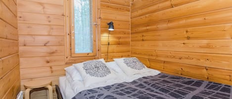 Una camera in legno con un letto, una finestra con persiane, uno sgabello e una lampada fissata al muro.