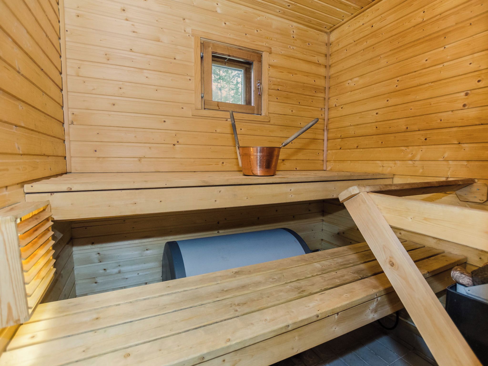 Ein Holzblockhaus-Innenraum mit Fenster, einem Metallkübel und einem blauen Gegenstand auf dem Boden.