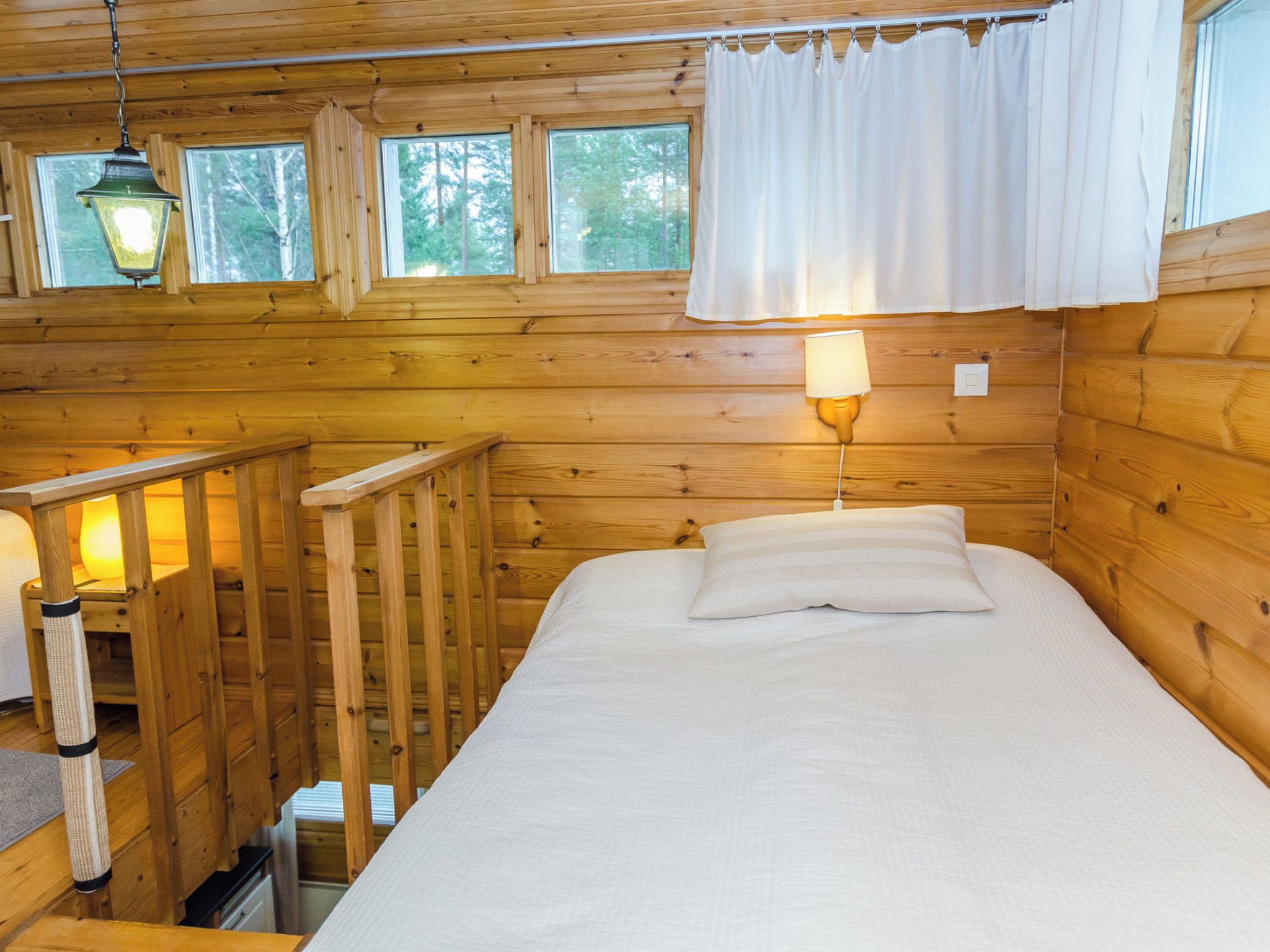 Habitación de cabaña de madera con una cama, una mesita de noche, una lámpara y una ventana con cortinas.