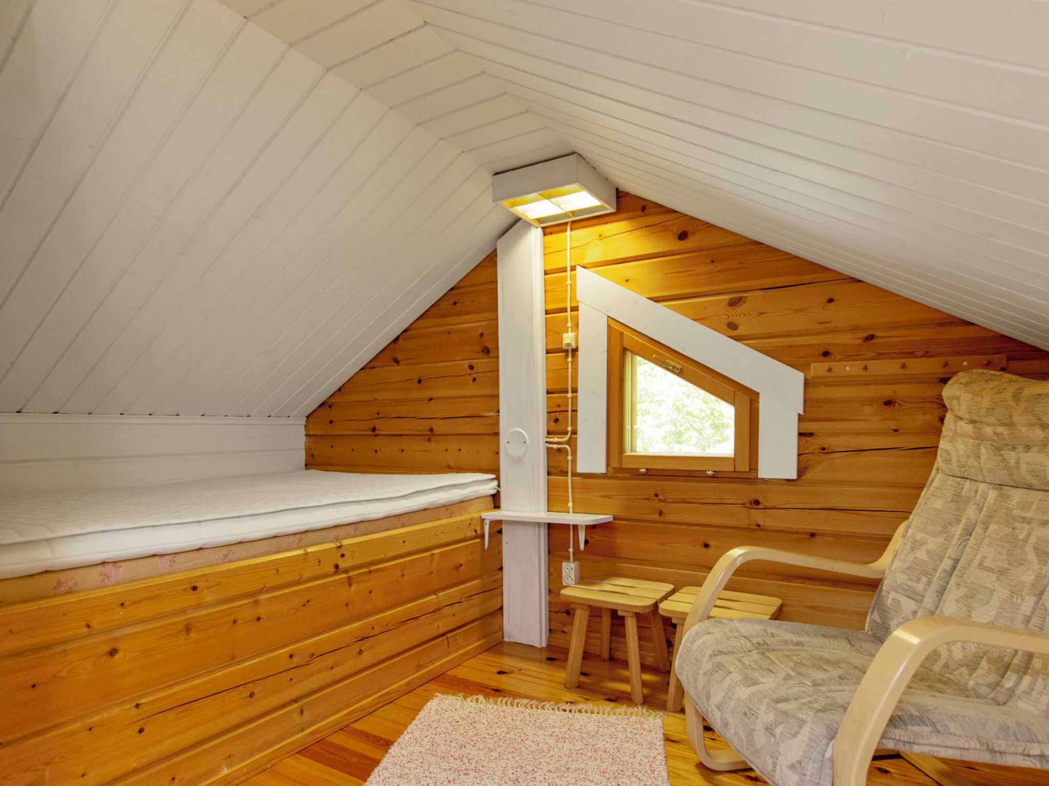 A cozy attic bedroom with wooden walls, a slanted ceiling, a small window, a bed, a chair, and a desk.