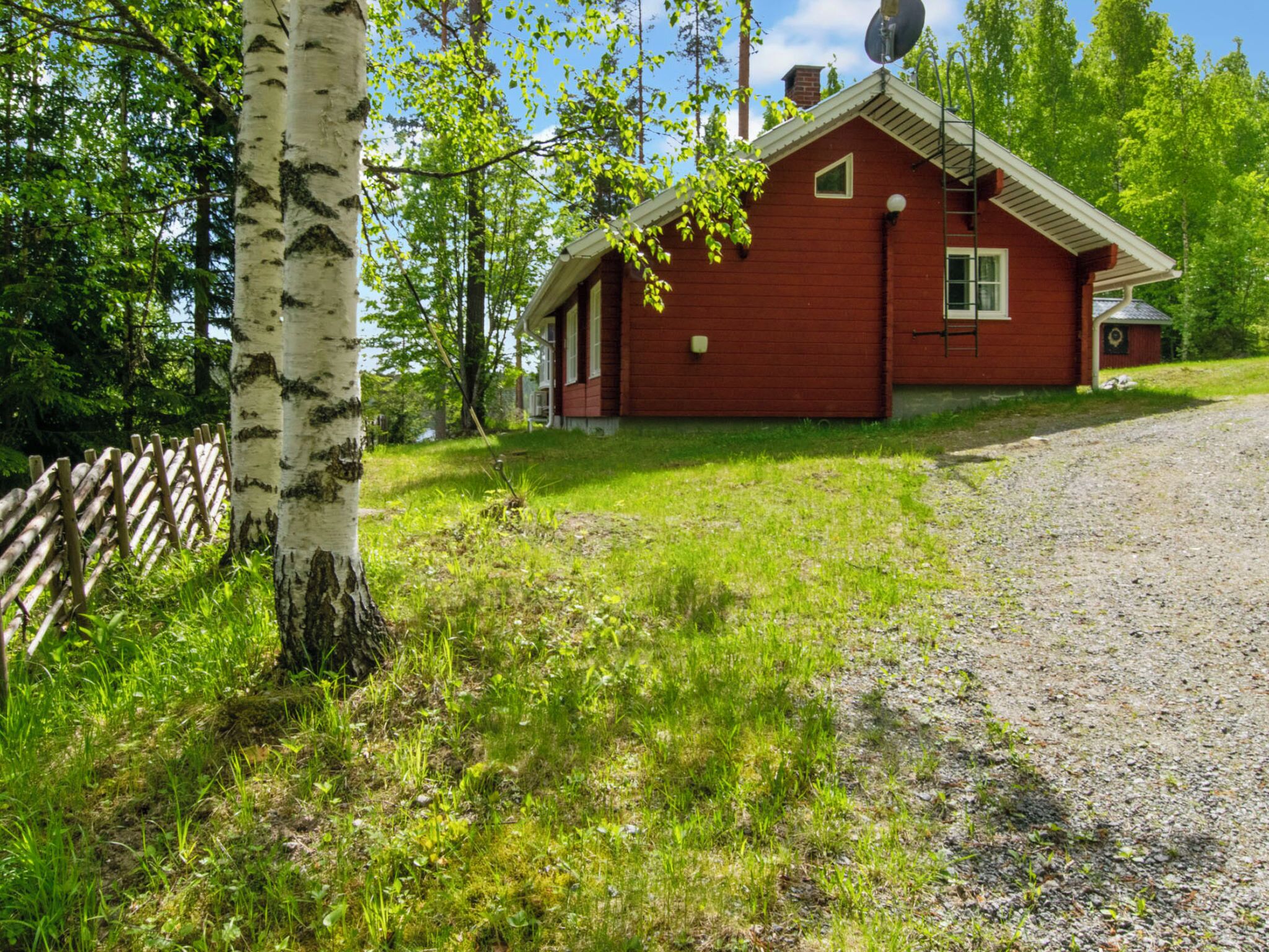 Punainen puinen mökki, jossa on savupiippu, ja sen ympärillä on puita sekä soralla päällystetty polku.