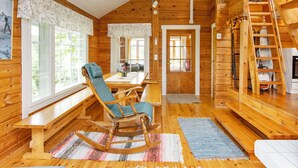 Ein gemütliches Blockhaus-Interieur mit Holzwänden, einem Essbereich mit Tisch und Stühlen, einem Schaukelstuhl, einer Treppe und einem großen Fenster.