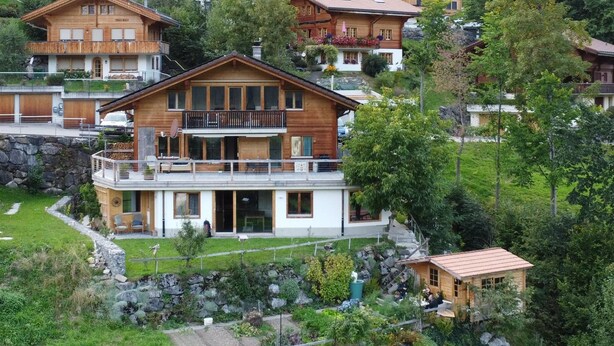 A large wooden chalet with a balcony, surrounded by lush greenery and other similar chalets in a mountainous area. - Strubelblick EG by Interhome (Lenk)