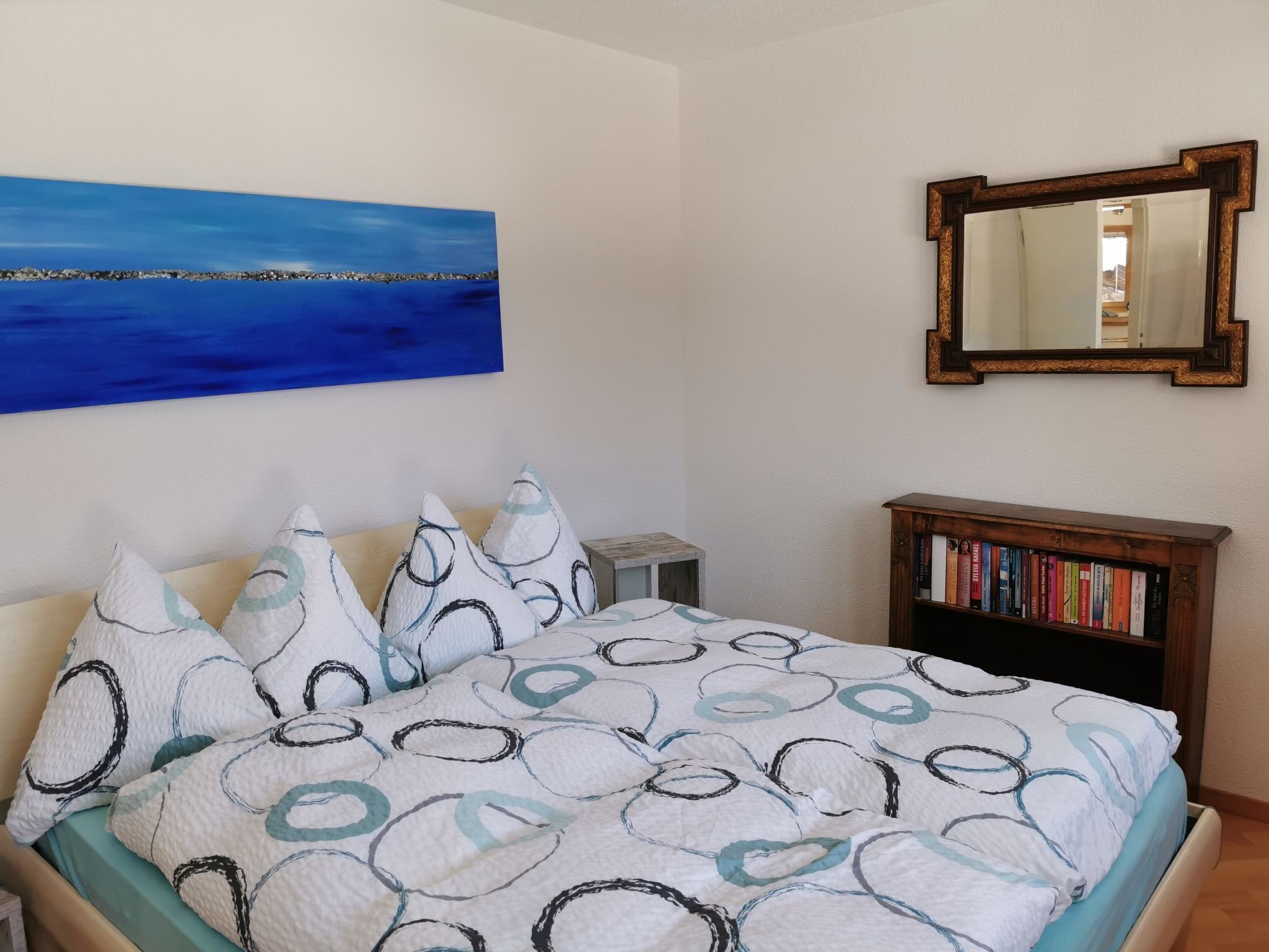 Une chambre à coucher avec un lit, des oreillers, un tableau, un miroir et une bibliothèque.