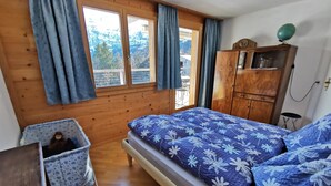 A bedroom with a wooden floor, a bed with a floral duvet, a wooden wardrobe, a small table, and a window with curtains. - Strubelblick EG by Interhome (Lenk)