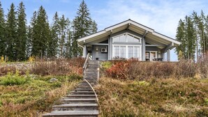 Een modern huis met een houten trap die naar de ingang leidt, gelegen in een natuurrijk landschap met bomen en struiken.
