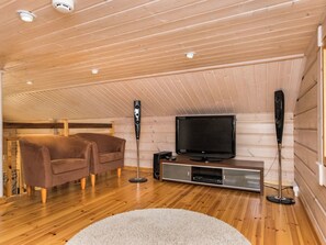Een gezellige kamer met houten vloeren, een flatscreen televisie op een houten standaard, twee bruine fauteuils, een witte vloerkleed en een plafond met houten balken en inbouwverlichting.