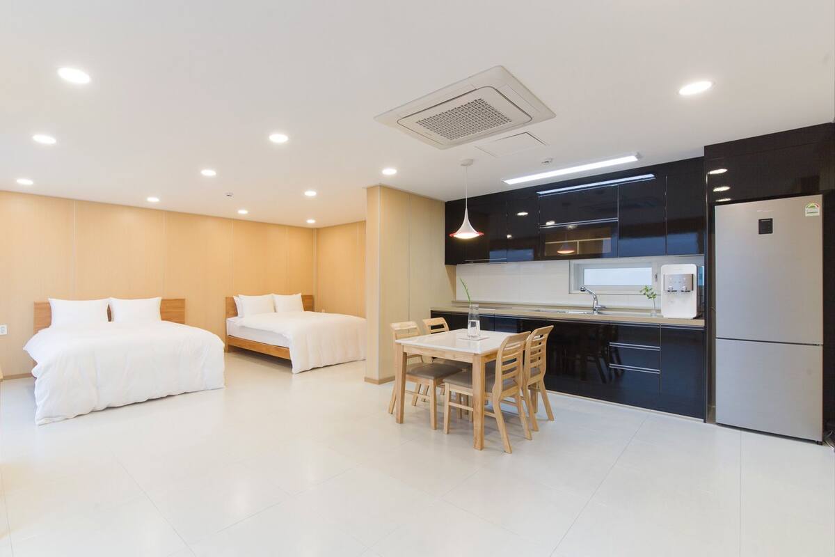 A modern hotel room with two beds, a dining area, and a kitchenette.