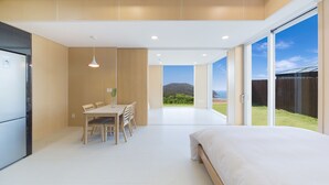 A modern bedroom with a large bed, a dining area with a table and chairs, and a view of a beach and mountains through large sliding doors. - The Mare Pool Villa Pension (Taean)