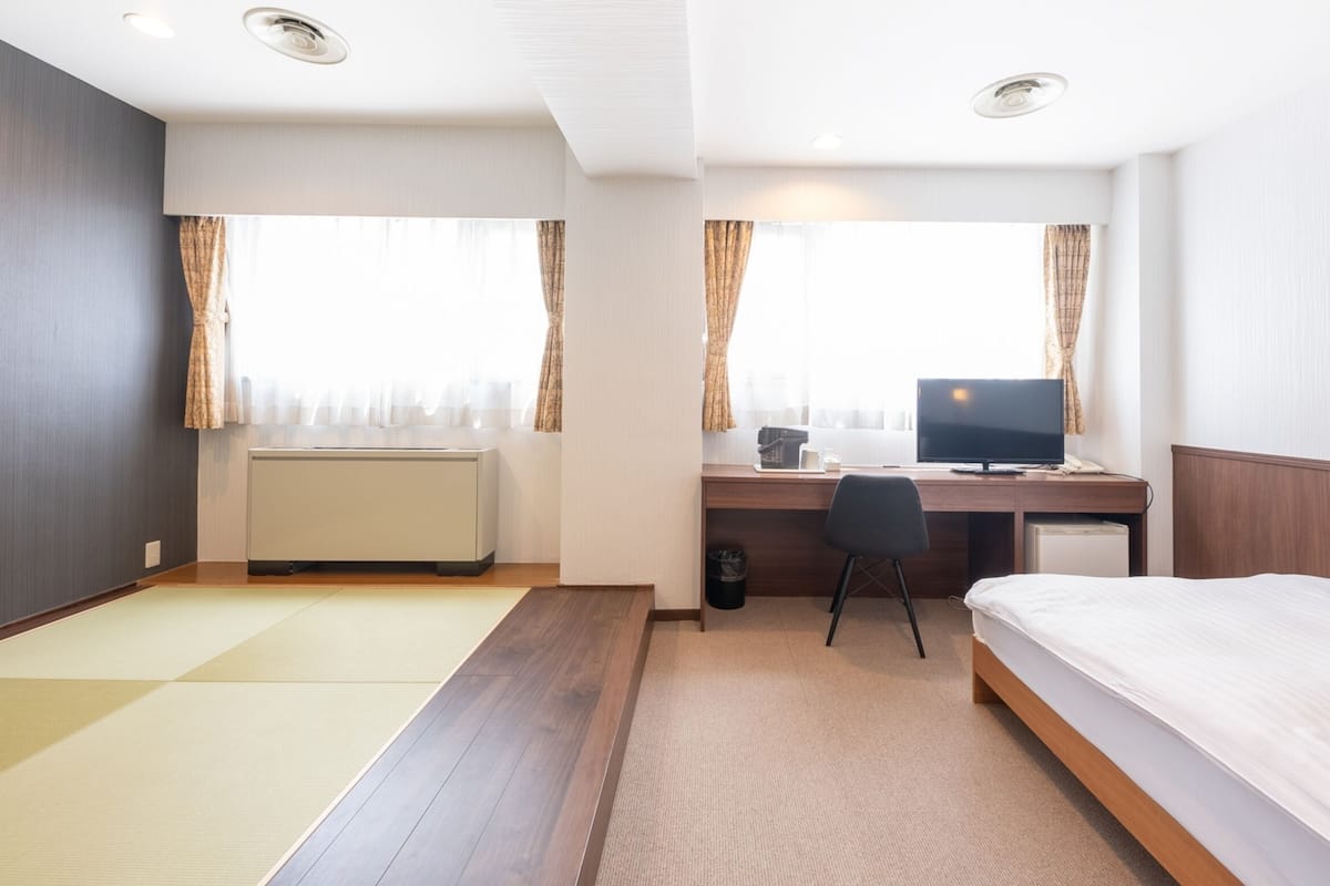 A hotel room with a bed, a desk with a computer, a chair, a radiator, and a window with curtains.