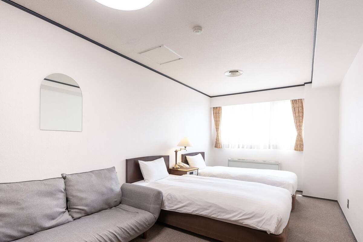 A hotel room with a grey sofa, two single beds, a bedside table with a lamp, a mirror, and a window with curtains.