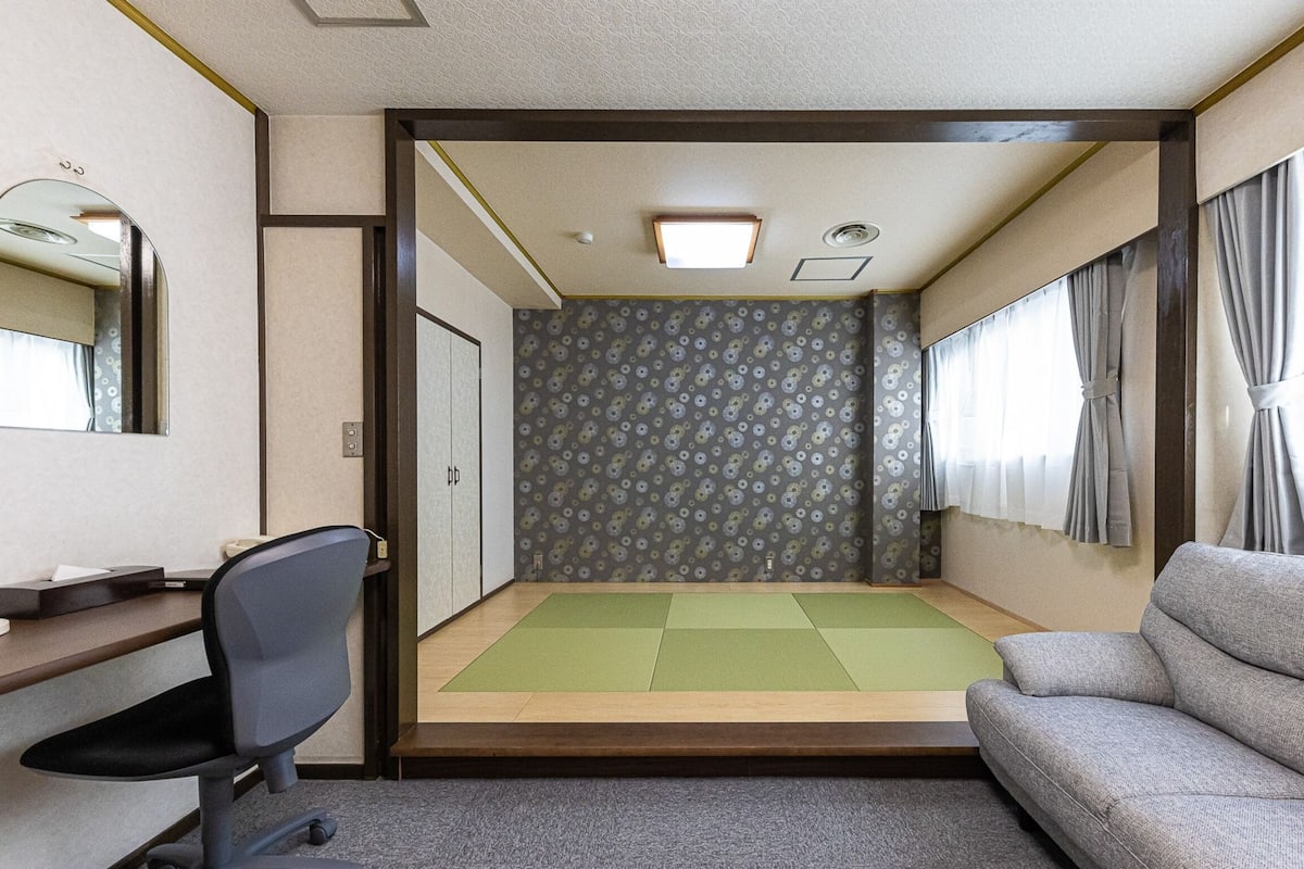 A compact room with a desk, chair, sofa, and a small tatami mat area.