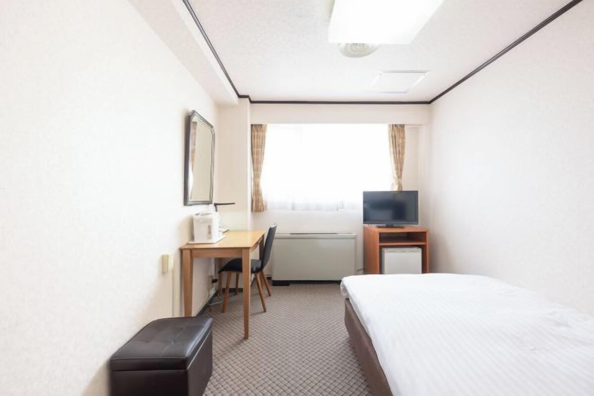 A hotel room with a bed, a desk with a chair, a television, and a window with curtains.
